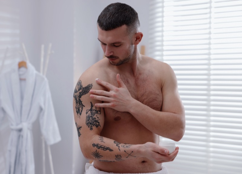 Man with tattoos in the bathroom putting cream on his shoulder