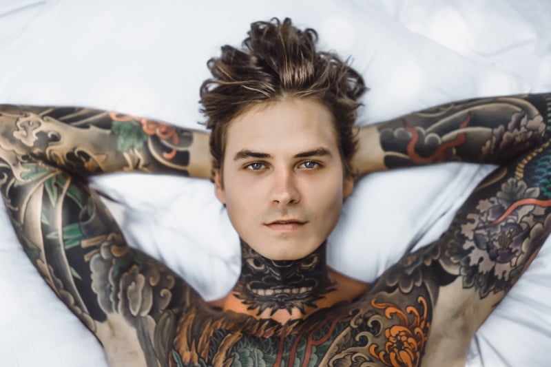 A man with tattoos lays in bed looking directly at the camera