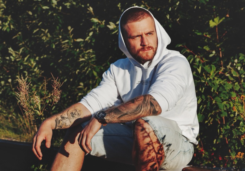 A man with tattoo sits outside wearing a white hoodie and denim shorts