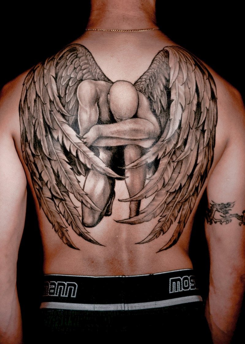 Black and gray fallen angel tattoo spanning a man's entire back, depicting a kneeling winged figure with large feathered wings from shoulder to hip.