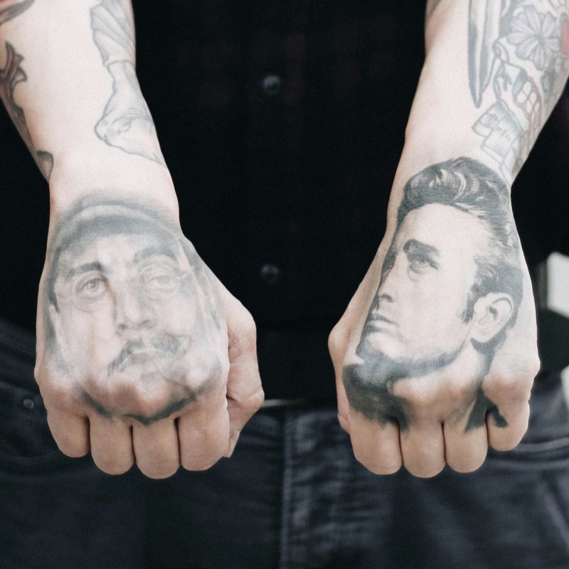 Two clenched fists held forward, each displaying a black-and-grey portrait tattoo across the back of the hand — a mustachioed man on one and a slick-haired James Dean-style face on the other.
