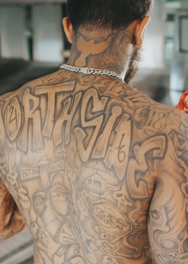 Full back tattoo in black and gray Chicano-style lettering and imagery on darker skin, with bold text and portrait elements covering the entire back.