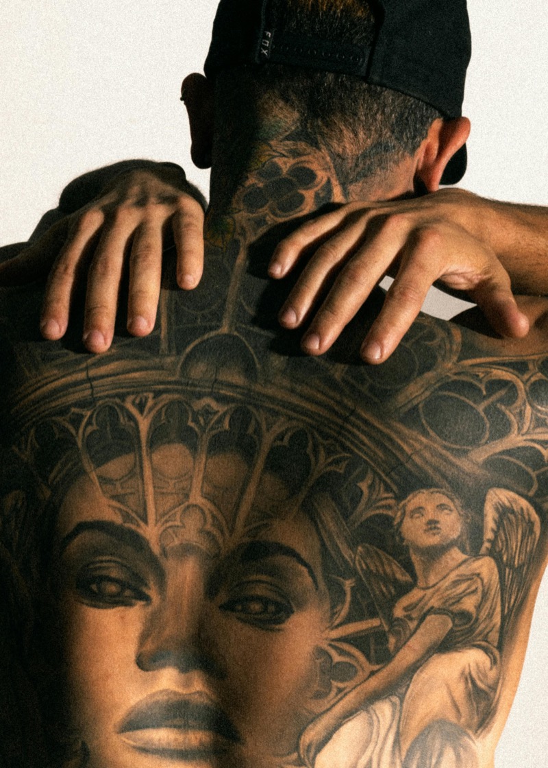 Man reaching behind his neck to reveal a full back piece in black and gray realism showing a woman's portrait and Gothic cathedral architecture.