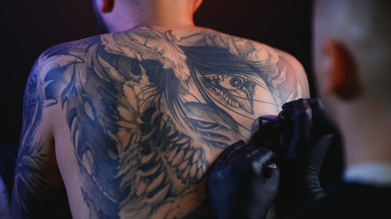 Tattoo artist in black gloves working on a large black and gray realism back piece, showing a detailed eye design surrounded by feathers mid-session.