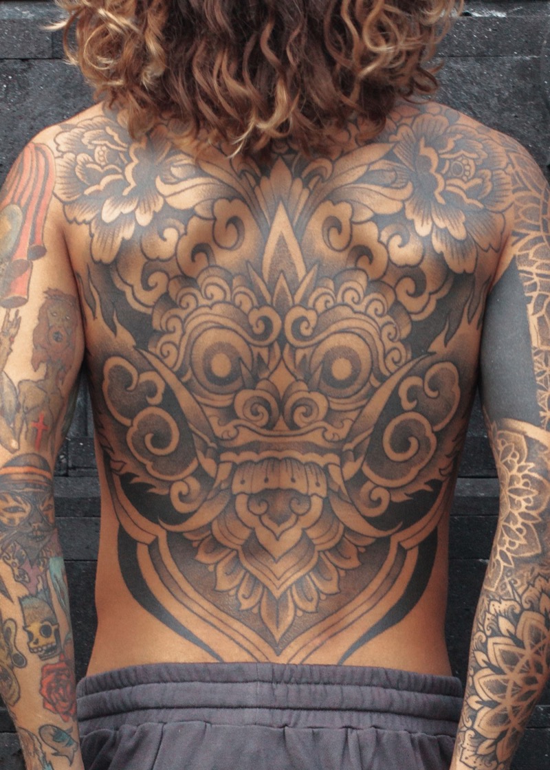 Full back tattoo of a Balinese Barong mask with ornamental scrollwork and floral patterns in black and gray, covering a man's entire back.