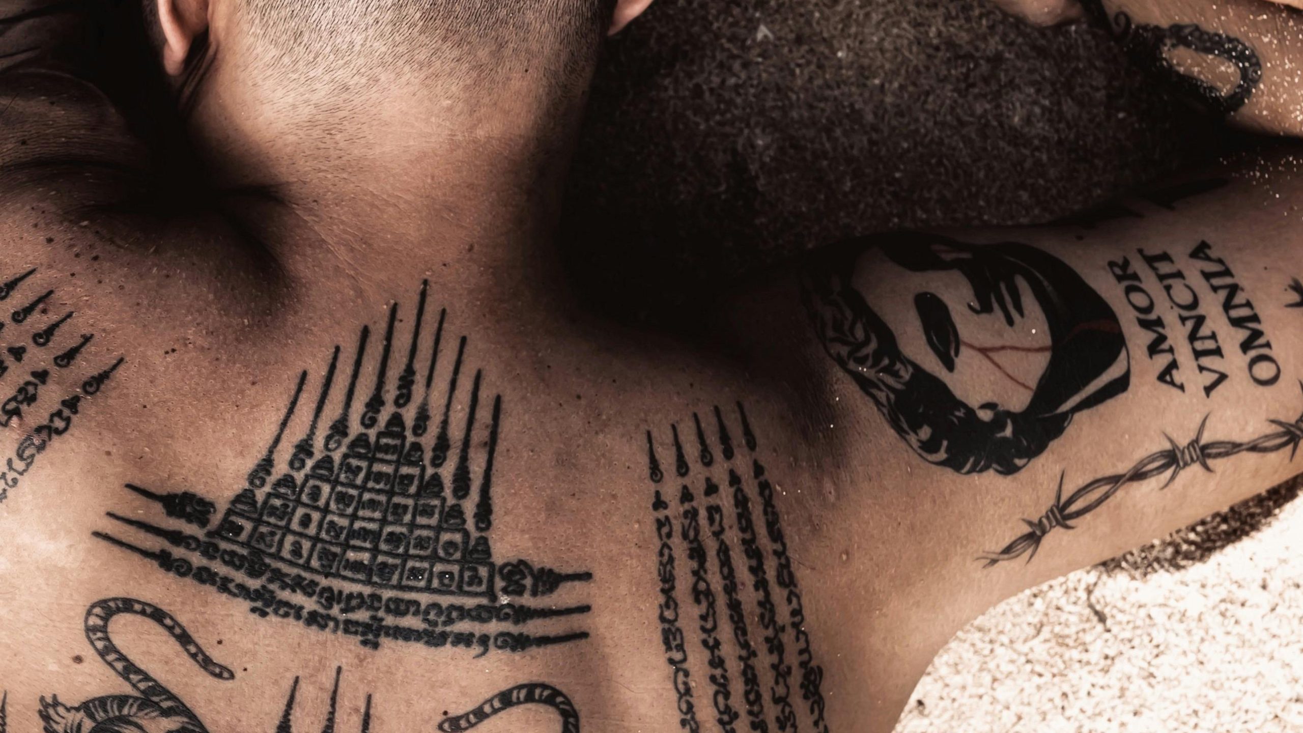 Man lying face down on sand with Sak Yant traditional Thai tattoos across his upper back, including sacred geometric patterns and script in black ink.