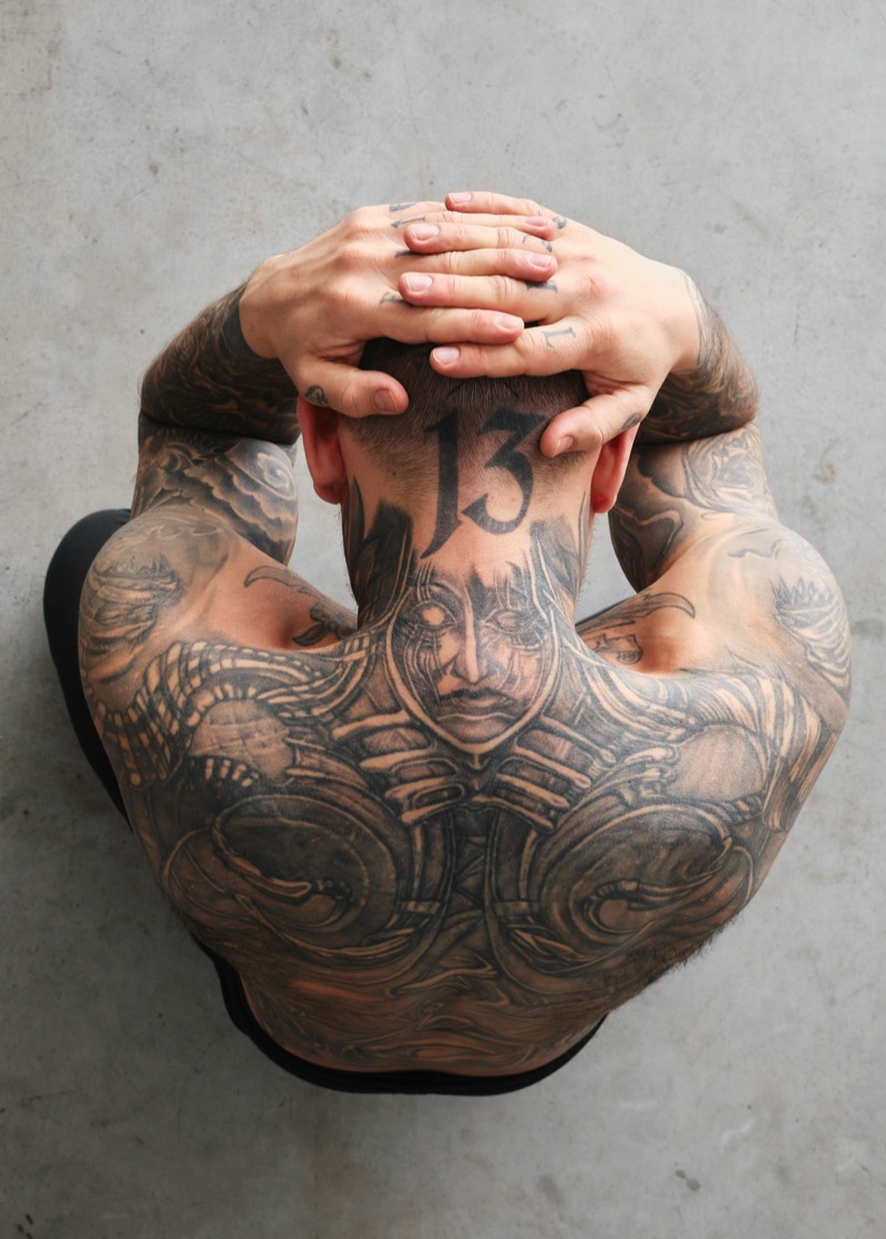Overhead view of a heavily tattooed man with hands behind his head, showing a full black and gray back piece flowing into both sleeve tattoos across the shoulders.