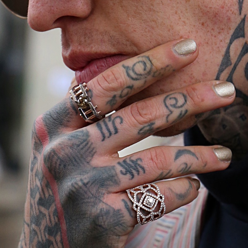 A close-up of a heavily tattooed hand near the mouth, with intricate dark designs flowing across the back of the hand, the fingers, and the webbing between them, accented by silver rings.