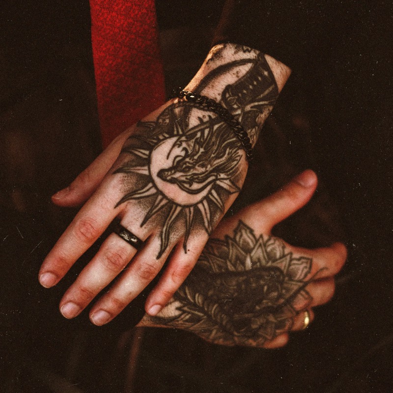 Two heavily tattooed hands clasped together, with a blackwork sun face on the back of one hand and a dense ornamental mandala-style design on the other.