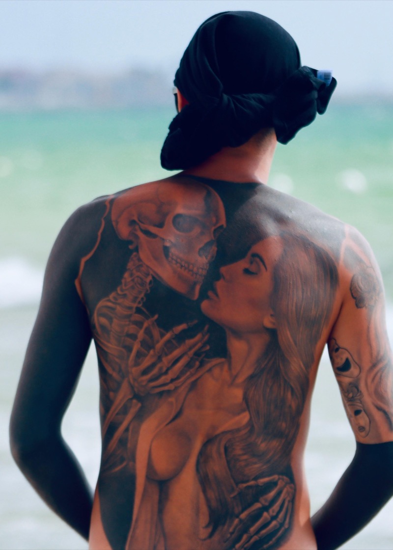 Black and gray realism full back piece showing a skeleton embracing a woman, covering the entire back with heavy shading, photographed at the beach.