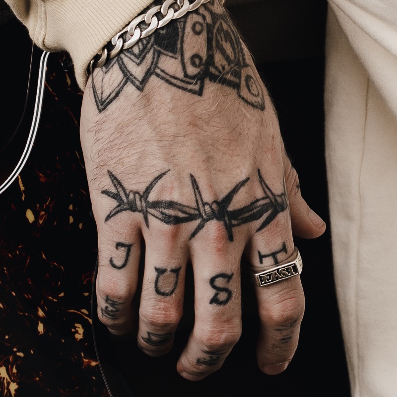 A bold barbed-wire tattoo wrapping across the knuckles of a man's hand, with block-letter knuckle ink and a silver ring.