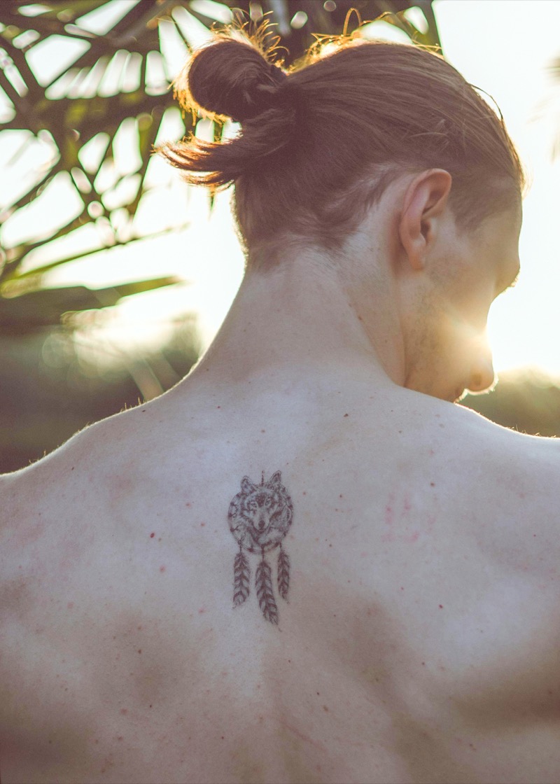 Small fine-line wolf and dreamcatcher tattoo centered between a man's shoulder blades, backlit by sunlight.