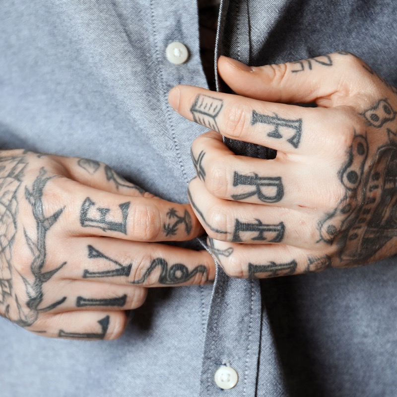 Two heavily tattooed hands resting against a grey button-down shirt, with bold knuckle lettering reading 'LIVE FREE' across both sets of fingers and small symbols on each digit.