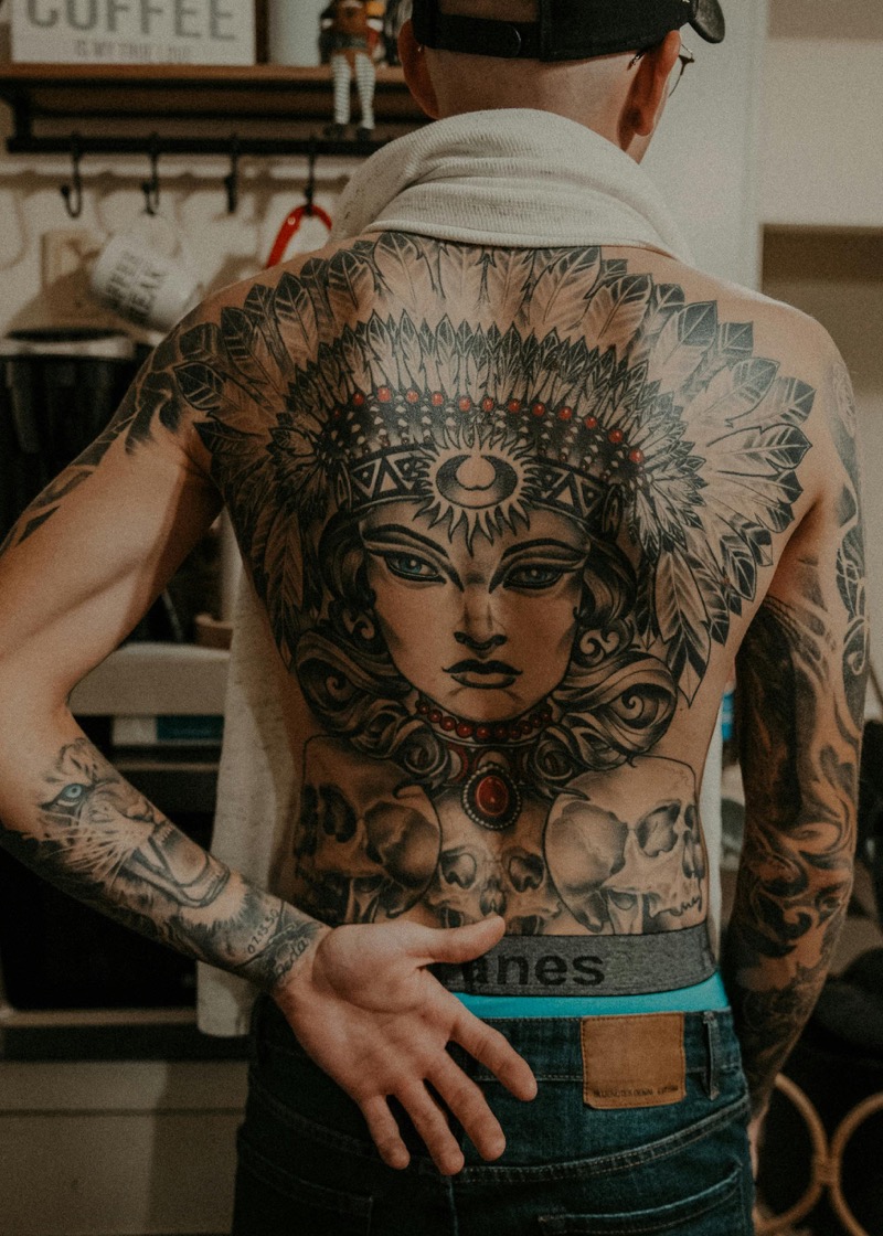 Full back piece in black and grey showing a large portrait with feathered headdress, skulls at the base, and detailed shading from shoulders to waistline.
