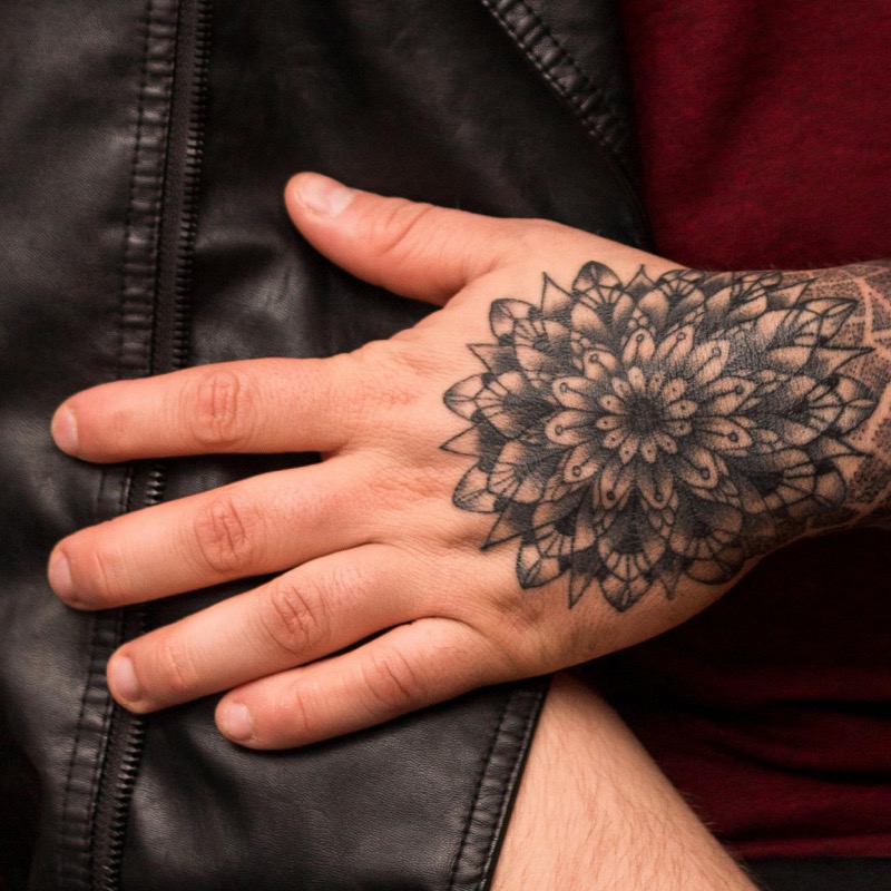 A detailed dotwork mandala tattoo with symmetrical geometric petals covering the back of a man's hand, set against a black leather jacket.
