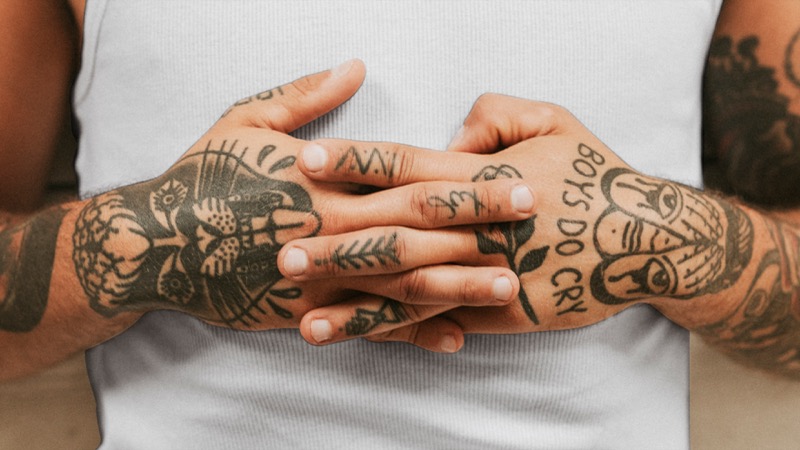 Two heavily tattooed hands clasped together in front of a white tank top, covered in traditional black ink including a panther face and 'BOYS DO CRY' lettering across the knuckles.