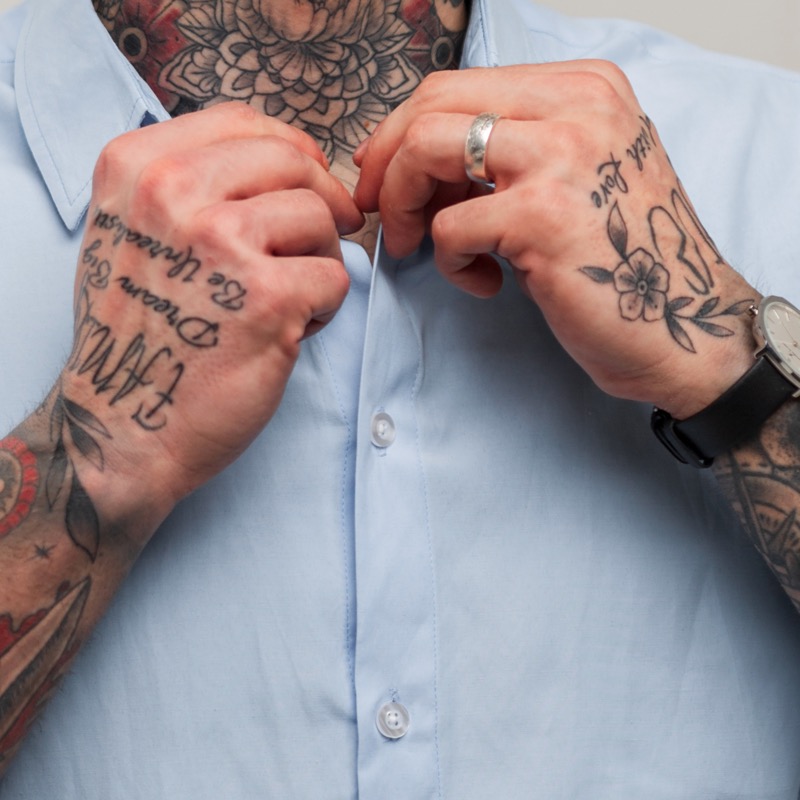 Two tattooed hands buttoning a light blue dress shirt, with cursive script across one hand and a flower-and-leaf tattoo across the other, alongside a watch and silver ring.