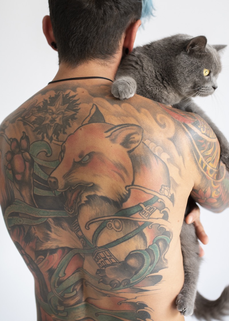 Man holding a gray cat over his shoulder, revealing a colorful neo-traditional full back tattoo of a fox with waves and floral elements in orange, green, and blue.