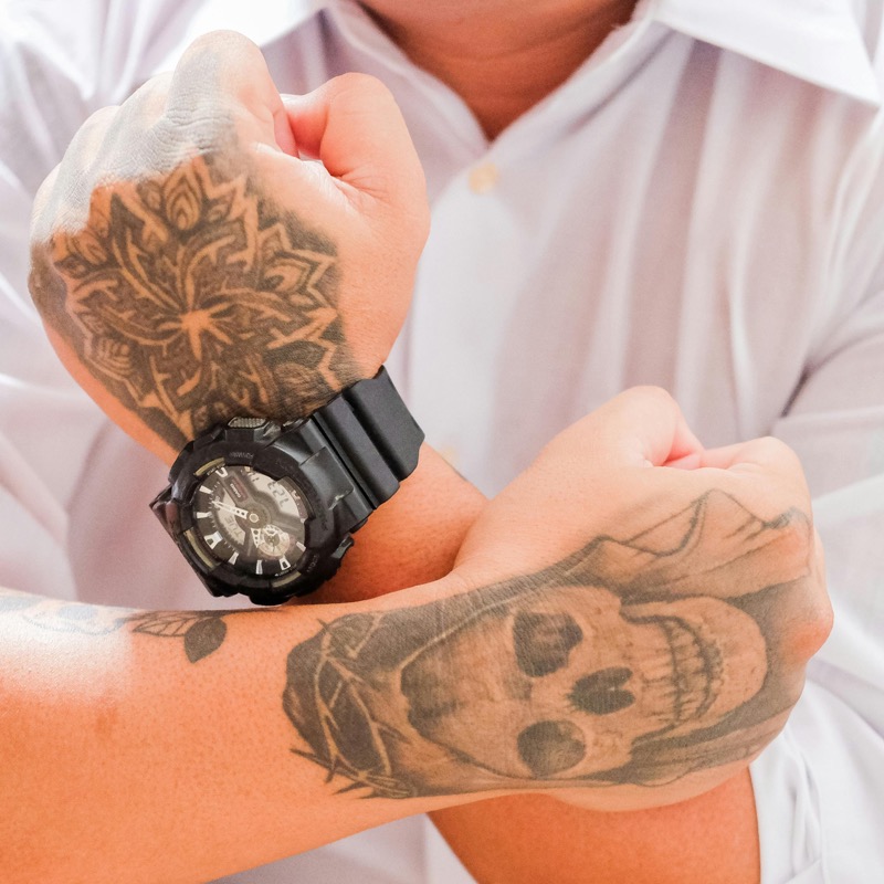 Crossed forearms showing a softened mandala tattoo on the back of one hand and a faded grey skull tattoo on the inside of the other wrist, with a black sports watch.