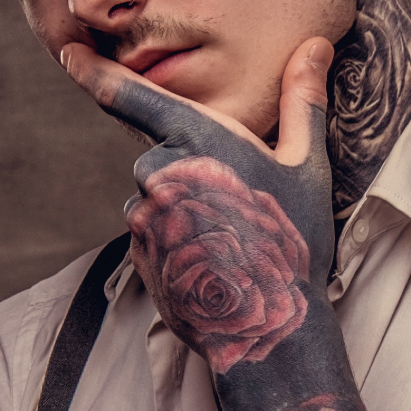 A man cradles his face with a tattooed hand featuring solid blackout shading and a realistic red rose tattoo layered over the dark fill, with more rose work climbing his neck.
