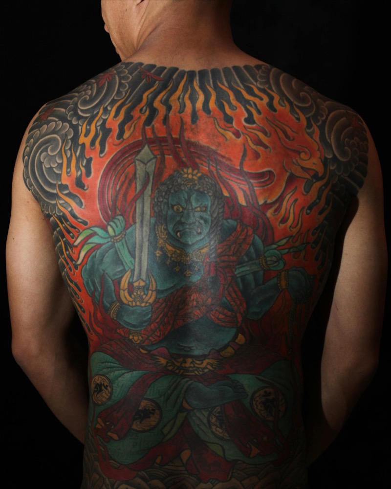 Traditional Japanese irezumi full back piece depicting Fudo Myoo in blue and green surrounded by red flames and gray cloud bars extending to both shoulders.