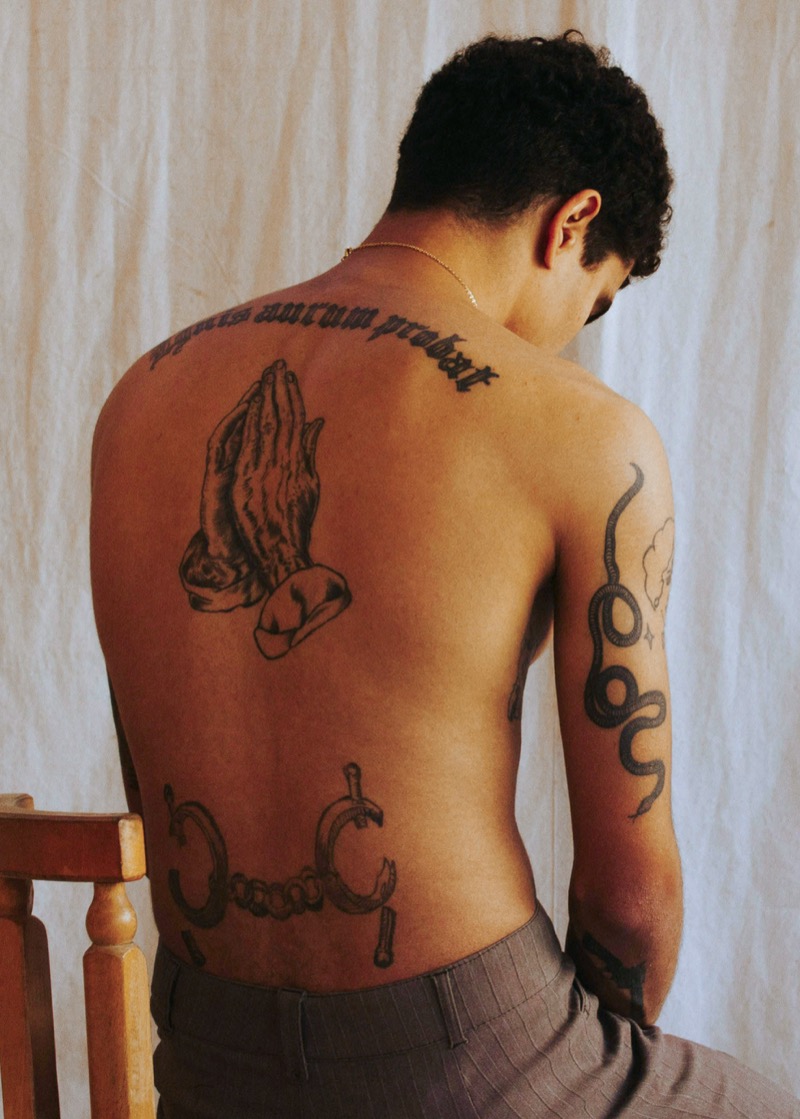 Man seated showing back tattoos including praying hands on the center back, Gothic script across the upper back, and chain links on the lower back.