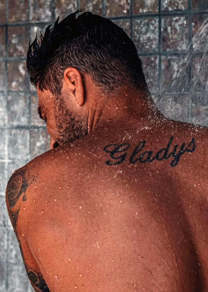 Man in a shower with the name Gladys tattooed in cursive script across his upper back between the shoulder blades.