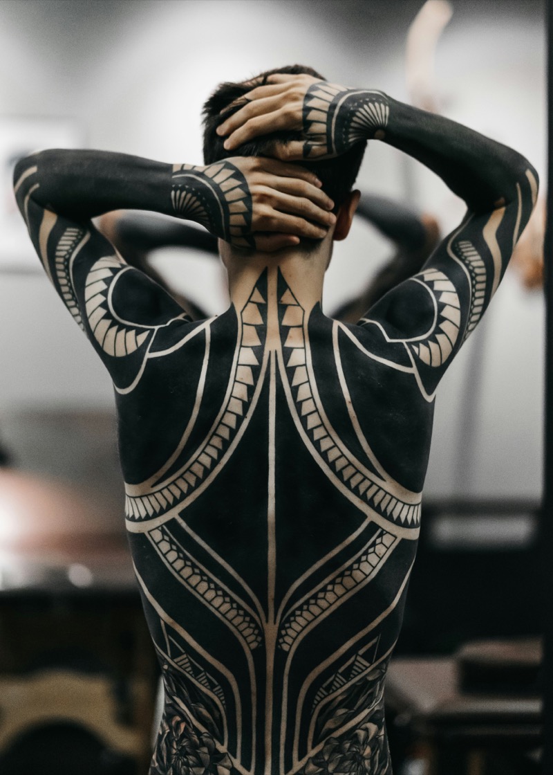 Full blackwork bodysuit covering a man's entire back and both arms, using negative space to create geometric and tribal patterns in solid black ink.