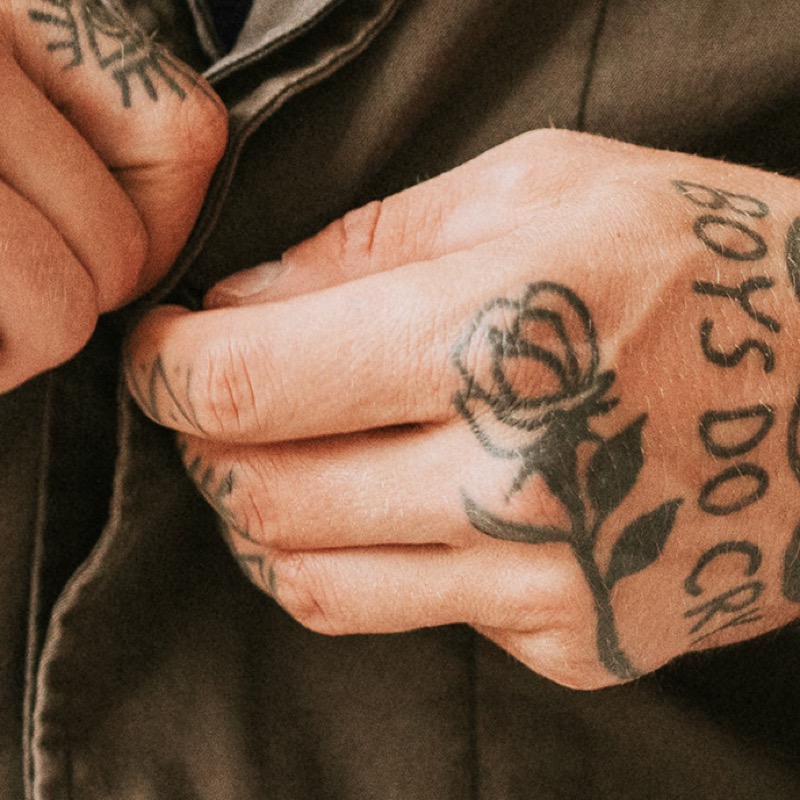 A man's tattooed hands buttoning a brown jacket, with a bold black rose outline on the back of one hand and 'BOYS DO CRY' lettering along the side of the hand and fingers.