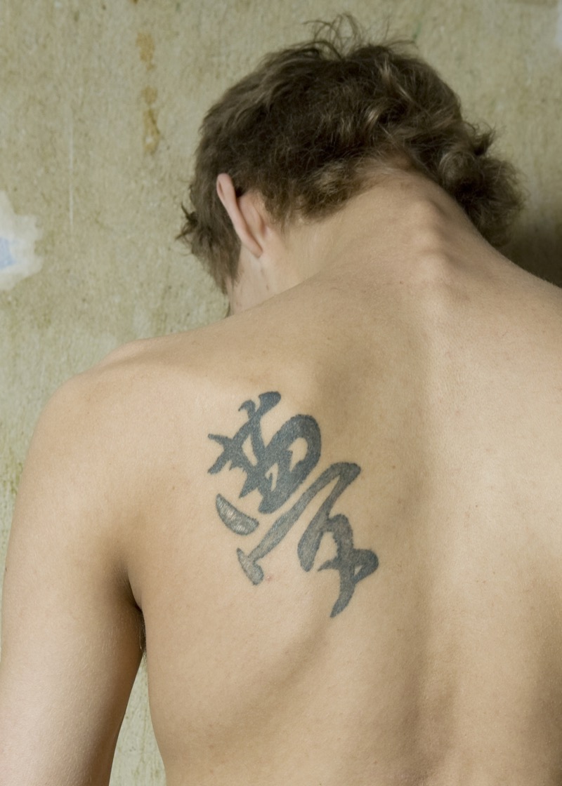 Faded calligraphic characters tattooed on a man's left shoulder blade in gray-black ink against bare skin.