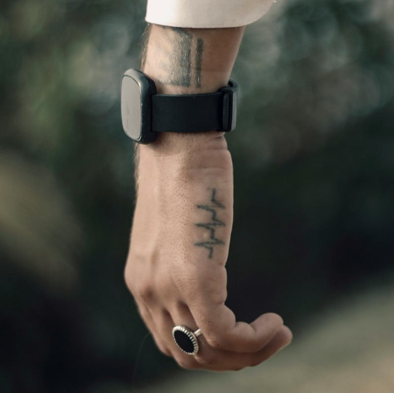 A simple heartbeat-line tattoo running along the side of a man's wrist and hand, with a black smartwatch and silver ring.