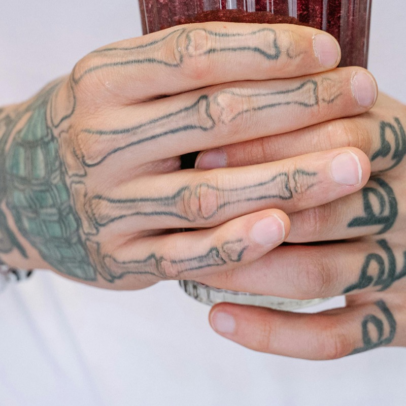 Two hands gripping a glass of red beverage, with a detailed skeleton hand tattoo in black ink mapping bone structure across the back of one hand and down each finger.