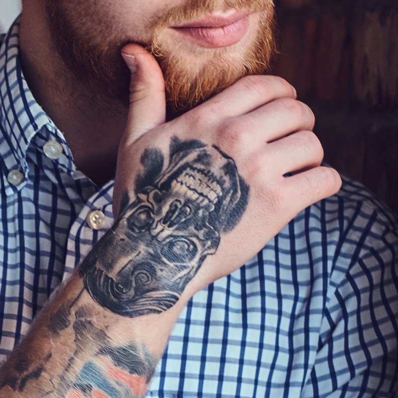 A man with a red beard rests his tattooed hand against his chin, showing a detailed black-and-grey skull tattoo across the back of the hand.