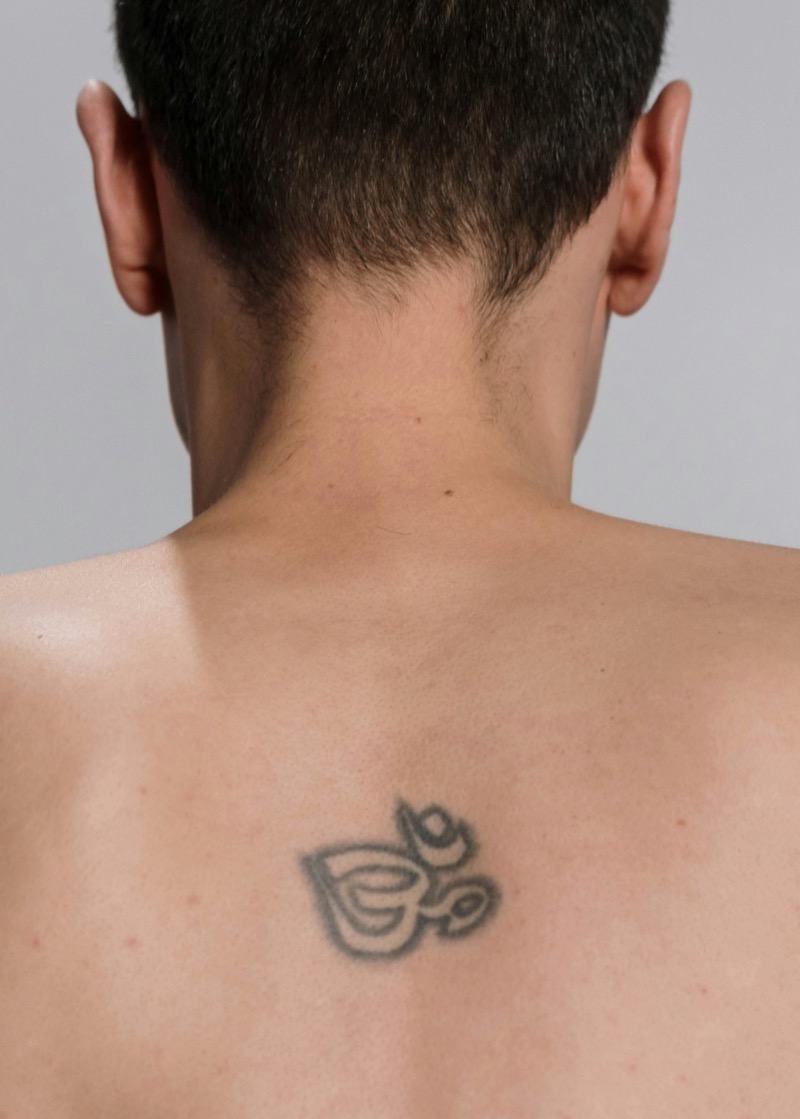 Small Om symbol tattoo in faded gray ink centered between a man's shoulder blades on otherwise bare skin.