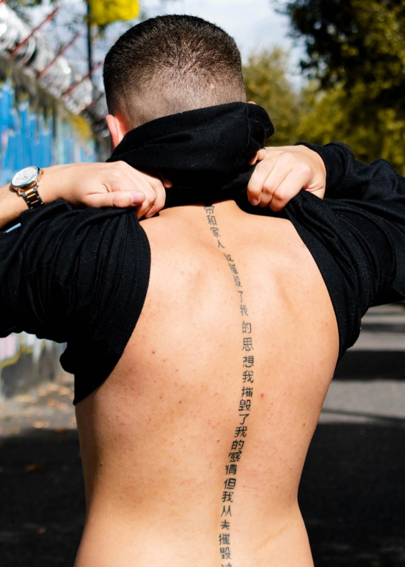 Man pulling up his black hoodie to reveal Chinese characters tattooed vertically down the center of his spine.
