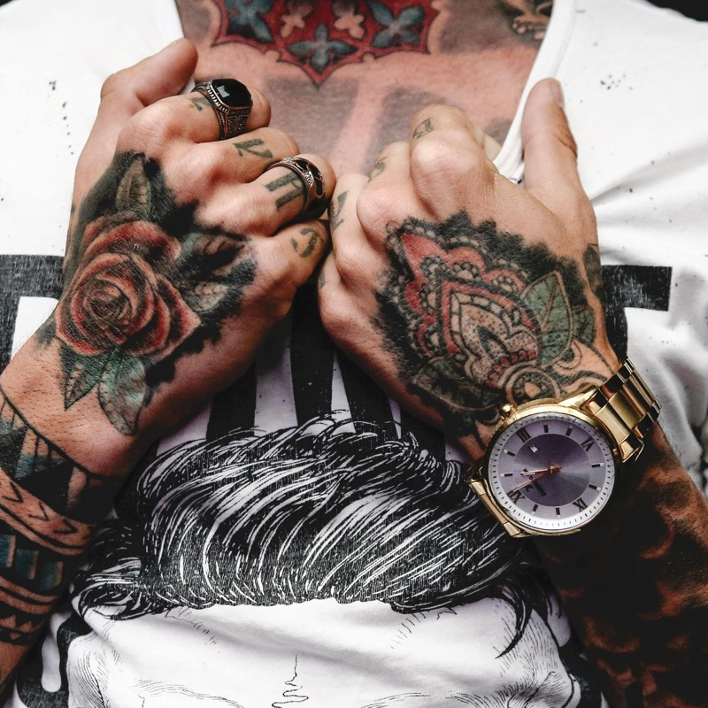 Two heavily tattooed hands clasped together over a printed white tank top, displaying a traditional black-and-red rose on one hand and an ornamental design on the other, with a gold watch on the wrist.
