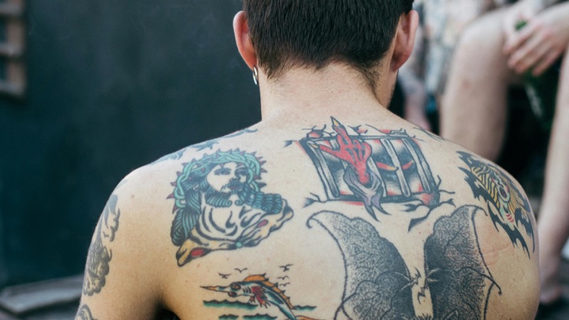 Man's upper back covered in American traditional tattoos including a Jesus portrait, red devil hand, and swordfish in bold outlines with saturated color.