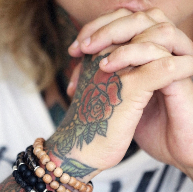 Two hands clasped together with a traditional rose tattoo in saturated red and green ink wrapping the back of the wrist and hand, alongside layered wooden-bead bracelets.