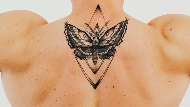 Black and gray moth tattoo framed by geometric diamond shapes centered on a man's upper back between the shoulder blades.