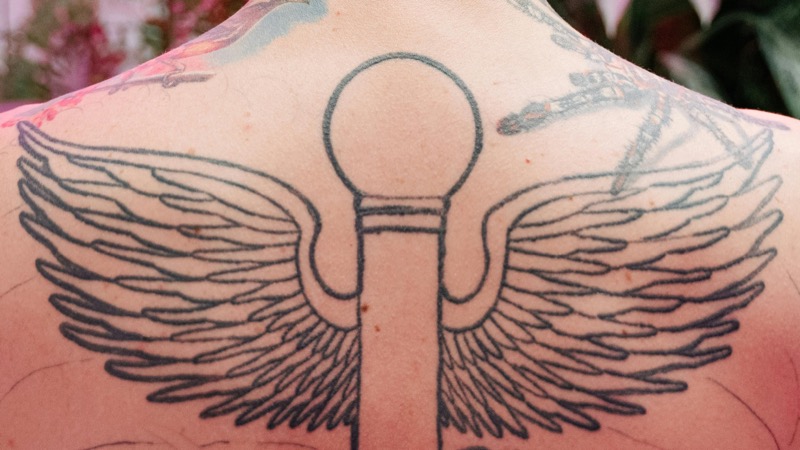 Winged caduceus tattoo spanning a man's upper back between the shoulder blades, with feathered wings extending outward in black ink.